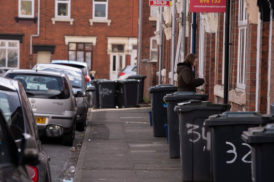 A woman standing on a residential sidewalk next to a row of large black wheelie bins with closed lids, placed along a brick building facade. The bins are used for rubbish collection and are positioned on the concrete pavement, which has visible wear and scattered debris. The brick building features multiple windows with white frames, and a red sign hanging above indicating property sales. In the background, several parked cars occupy the street, with some angled towards the curb. The scene is lit by natural daylight, casting soft shadows, and the overall environment suggests a typical urban residential area where private waste collection services, such as those provided by Waste Collection Streatham, may be utilized for on-site rubbish disposal. The presence of graffiti on some bins and the general tidy yet functional setting reflect everyday waste management practices in a suburban environment.