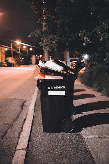A black wheeled rubbish bin labeled 'St. John's' is positioned on the pavement next to a curb in an urban street scene at night. The bin is partially open, revealing various waste items, including cardboard boxes and plastic packaging, stacked inside. The surrounding environment includes a dark, leafy hedge on the right, streetlights casting a warm glow, and a tree with dense foliage overhead. In the background, blurred streetlights and parked cars are visible, suggesting an evening scene in a residential or suburban area. The setting indicates an instance of private waste disposal or on-site rubbish collection, with the bin used for disposing of household waste which might be handled by independent waste collection services, such as Waste Collection Streatham. The overall atmosphere highlights a typical scene of waste awaiting collection, emphasizing the importance of proper rubbish management and disposal in local communities.