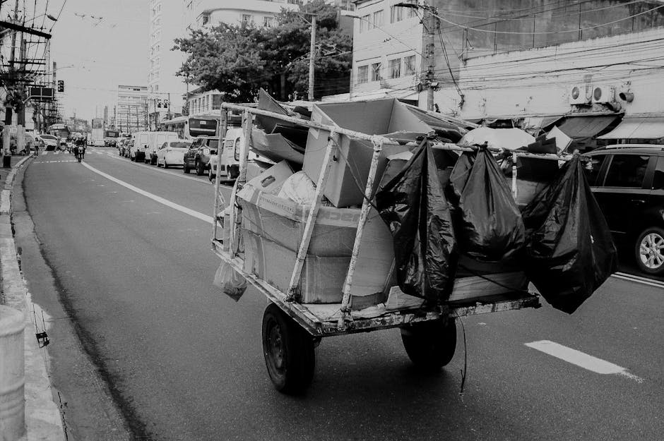 A small, wheeled metal cart loaded with various discarded items and rubbish bags, positioned on the edge of a city street. The cart contains large cardboard boxes, some partially crushed, and is secured with wooden supports and straps. Several black plastic rubbish bags are attached to the sides of the cart, appearing full and tied at the top. The street background includes parked cars lining the curb, multi-storey buildings with visible windows and external air conditioning units, and power lines overhead. The road surface is asphalt with painted white lane markings, and the scene is illuminated by natural daylight, casting subtle shadows on the street. This image depicts a scene related to private waste handling or alternative rubbish collection in an urban environment, consistent with rubbish removal services provided by companies like Waste Collection Streatham, aimed at small-scale or on-site rubbish clearance for businesses or residents.