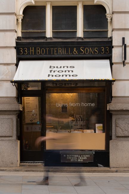 The image shows the exterior of a shop located at 33 H. Botterill & Sons, with a black and gold sign above the entrance displaying the address. The storefront features a white retractable awning with the partially visible text 'buns from home' printed on it, suggesting the shop specializes in baked goods or similar products. Behind the glass window, the interior is partially visible, revealing a countertop and some items that may be ingredients or packaging materials. The lower part of the window displays the text 'buns from home,' consistent with the awning, indicating the shop's branding. The building has a classic stone facade with decorative architectural elements framing the shop front, and the pavement in front appears clean with a neutral tone. Slight motion blur of a passing pedestrian is noted at the bottom of the image, emphasizing the busy street environment. This scene exemplifies an independent bakery storefront that might be serviced by private waste collection or rubbish removal services to manage packaging waste, bakery debris, or other refuse generated on-site.