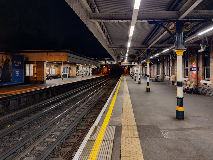 An empty platform at Streatham Hill station during nighttime, featuring illuminated overhead lighting and a row of cast-iron supporting columns with black and yellow striped accents. The platform surface is paved with concrete, including a tactile yellow warning strip along the edge. To the right, brick walls with several large windows and station signage are visible, while on the opposite side, there is a small building with a staircase and sheltered waiting area, illuminated by warm lighting. The railway tracks run parallel along the platform, extending into the distance and disappearing into the dark tunnel ahead. The scene is quiet, with minimal movement, reflecting a typical scene of an urban railway station prepared for passenger arrivals and departures, with clear indications of maintenance and structural elements that could involve routine cleaning or site management associated with station operations.